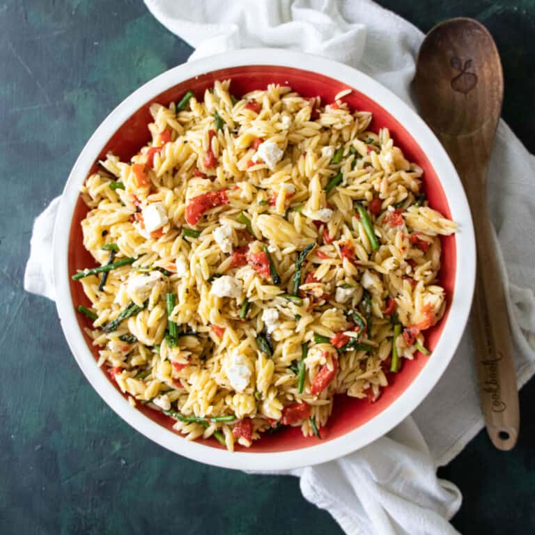 summer orzo salad in a serving bowl with a linen cloth and wooden spoon next to it.