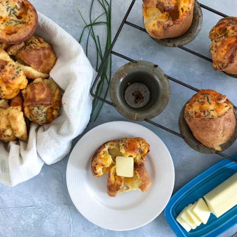cheddar and chive popovers on a blue background with butter, bread basket and muffins tin.