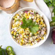 overhead shot of a white bowl of Healthy Mexican Street Corn Dip with salt, jalapeno, a linen and cilatro laying next to it on a blue background