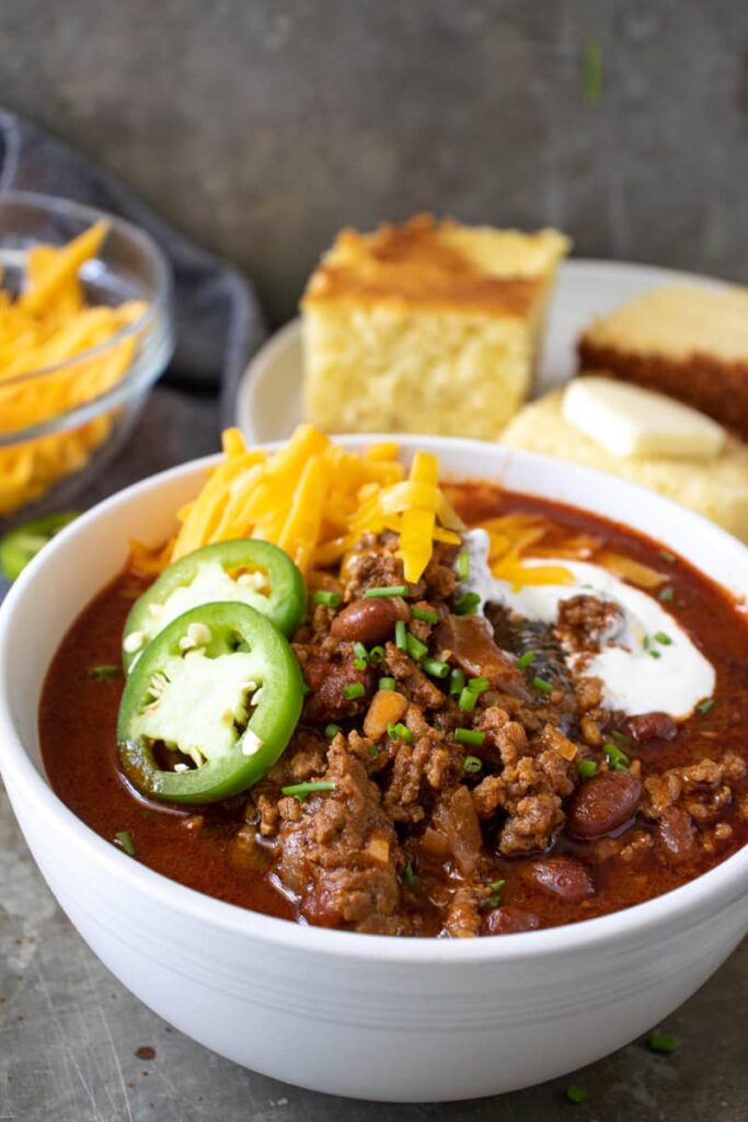 chili con carne in a bowl topped with cheese, sour cream, and sliced jalalpenos with cornbread in the background
