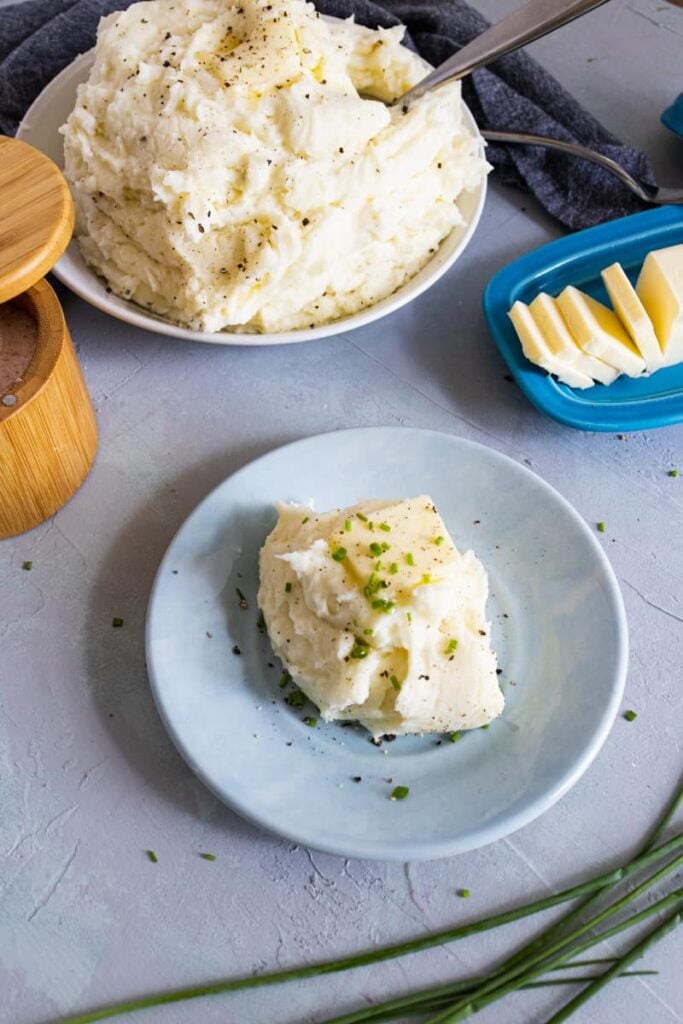 a blue plate with Creamy Southern Mashed Potatoes on it with a big bowl of potatoes in the back with butter, chives, and a wooden spoon next to it