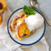 plate of fresh peach cobbler with a scoop of vanilla ice cream with a napkin, spoon, sliced peaches and mint