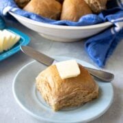 wheat dinner roll on a small blue plate with a pad of butter on top with a butter dish and bread bowl in the background