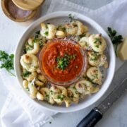 easy garlic knots in a white bowl with marinara sauce in the middle, a white napkin, parmesan a nd a salt container next to it