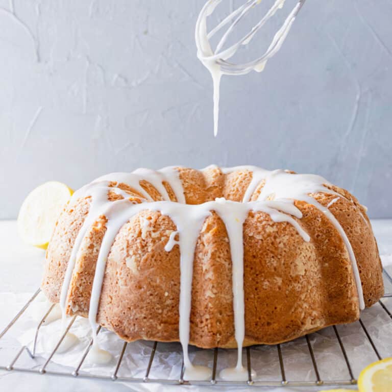 A whisk drizzling icing over a pound cake on a cooling rack.