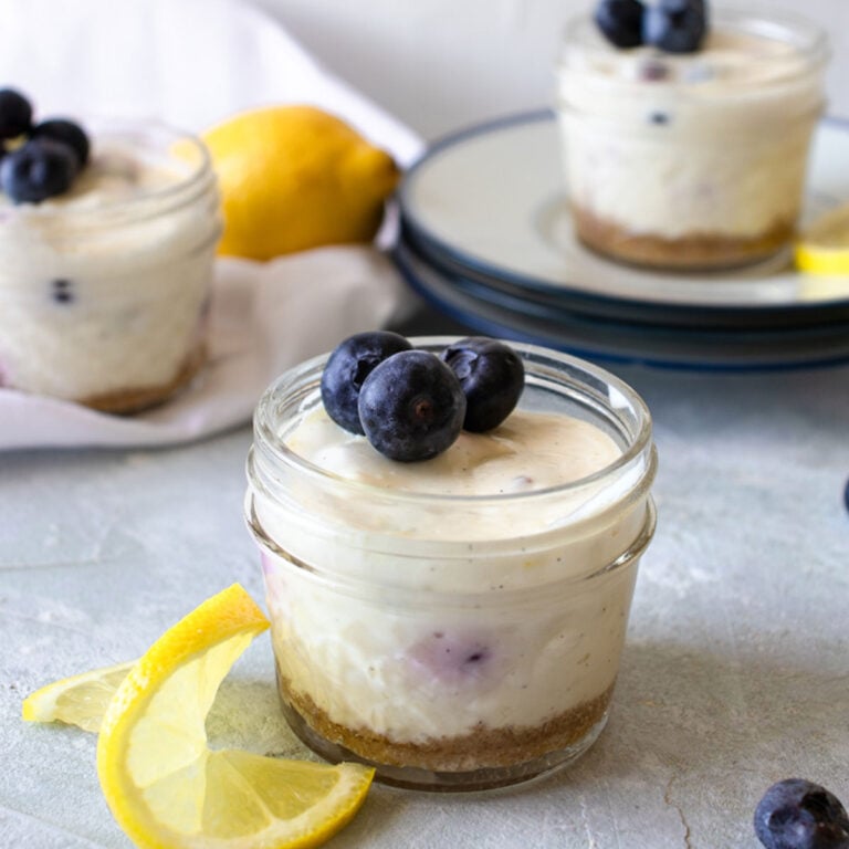 3 mason jars of lemon blueberry cheesecakes with lemon slices and blueberries next to them