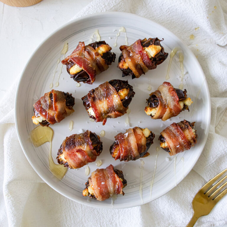 a white round platter of Goat Cheese Stuffed Dates Wrapped in Bacon drizzled in honey with a gold fork, salt, and napkin