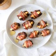 a white round platter of Goat Cheese Stuffed Dates Wrapped in Bacon drizzled in honey with a gold fork, salt, and napkin