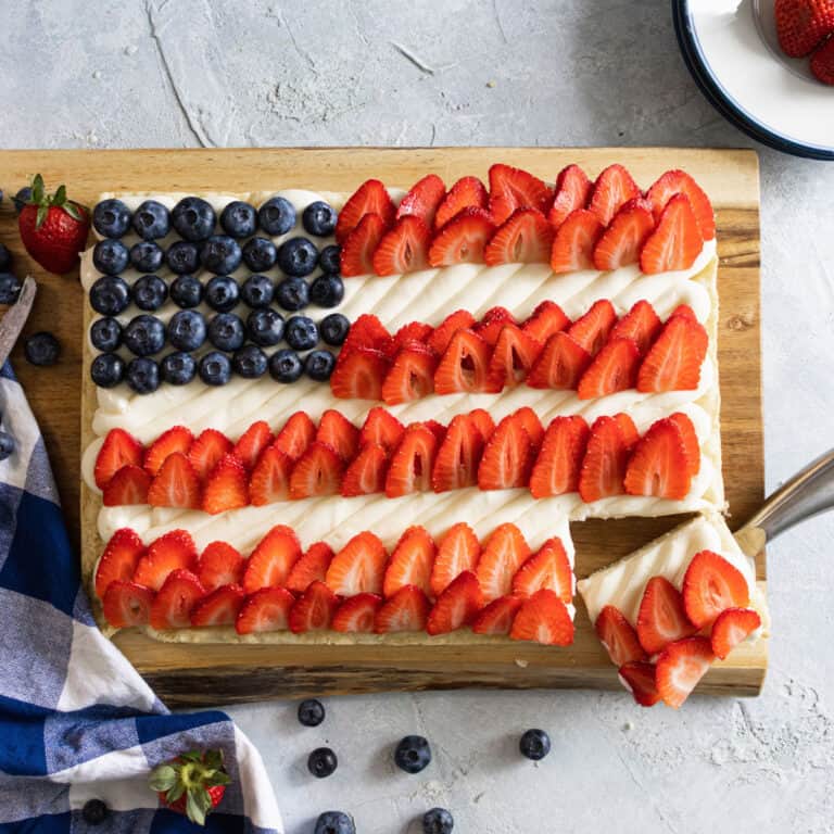 Red White and Blue Fruit Pizza ON A CUTTING board with a corner cut out. a napking, plate and berries beside it