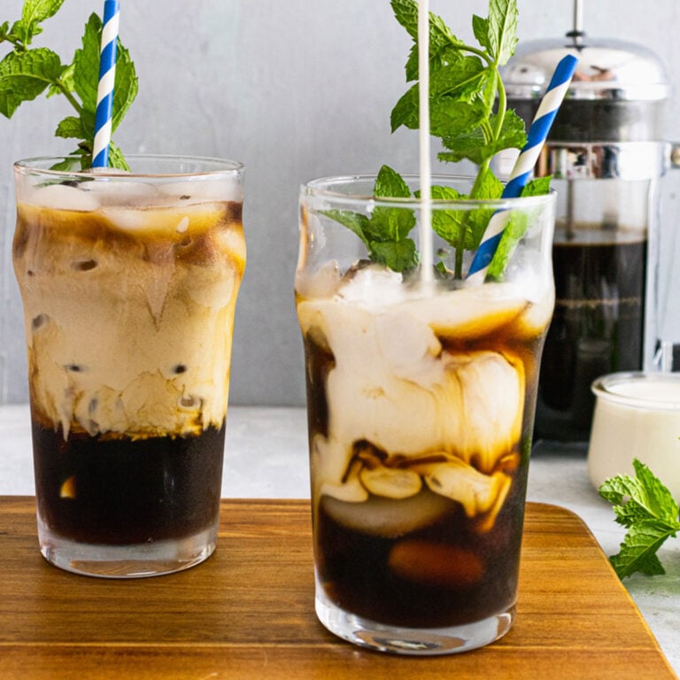 2 glasses of mint cold brew on a wooden cutting board with a glass of ice, french press and mint leaves in the background