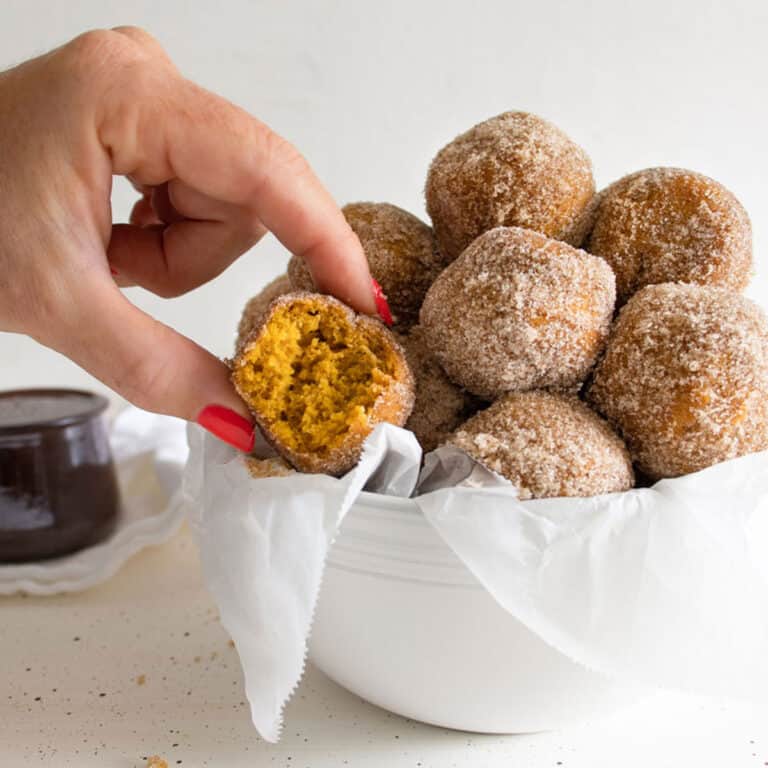 A hand holding a donut hole that has a bite taken out in a bowl of them. Chocolate dipping sauce in the background.