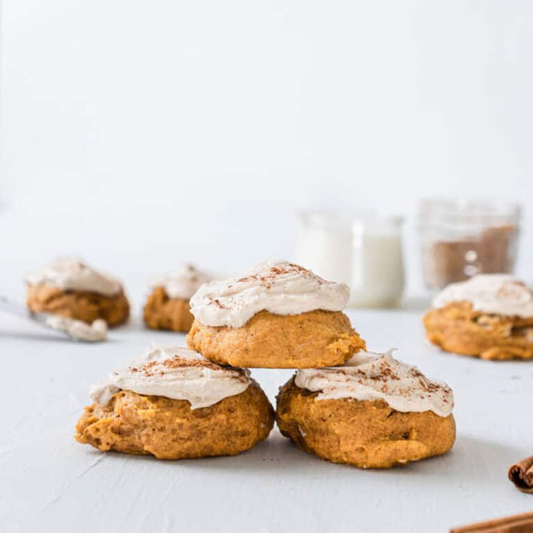 3 pumpkin cookies with brown butter icing stacked in a triangle with 3 cinnamon sticks in the foreground.