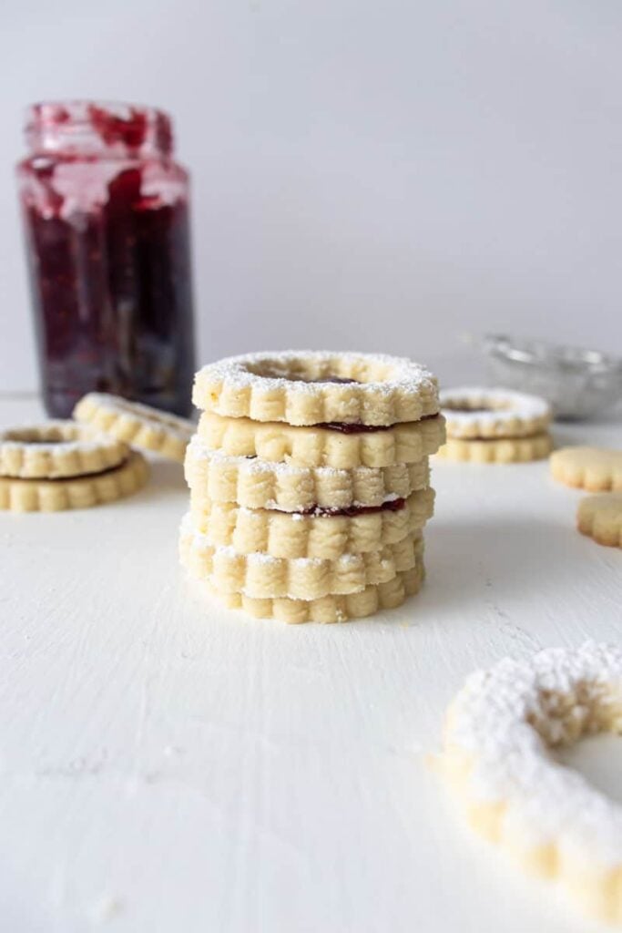 3 Shortbread Linzer Cookies stacked on top of each other with a jar of raspberry jam in the background