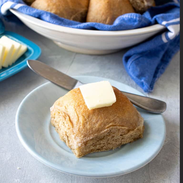 wheat dinner roll on a small blue plate with a pad of butter on top with a butter dish and bread bowl in the background