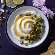 whipped feta dip with honey and pistachios on a purple table with lemons, crackers, cucumbers, and flowers next to the bowl.