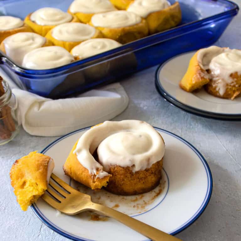A cinnamon roll on a plate with a small bite on a fork resting to the side. The pan of cinnamon rolls is in the background behind it.