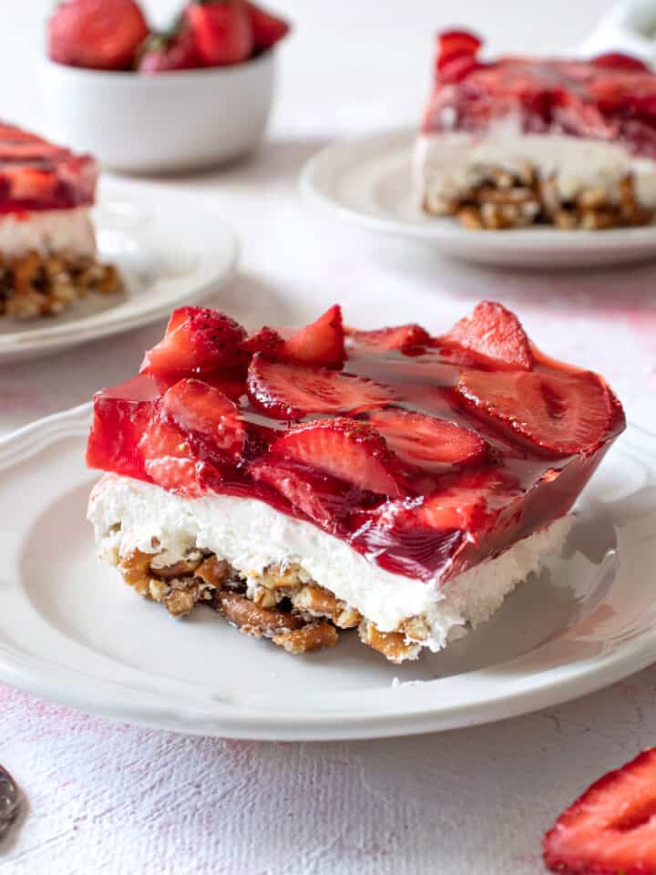 A slice of strawberry pretzel jello salad on a small white plate with 2 others in the background.