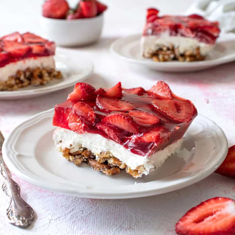 A slice of strawberry pretzel jello salad on a small white plate with 2 others in the background.