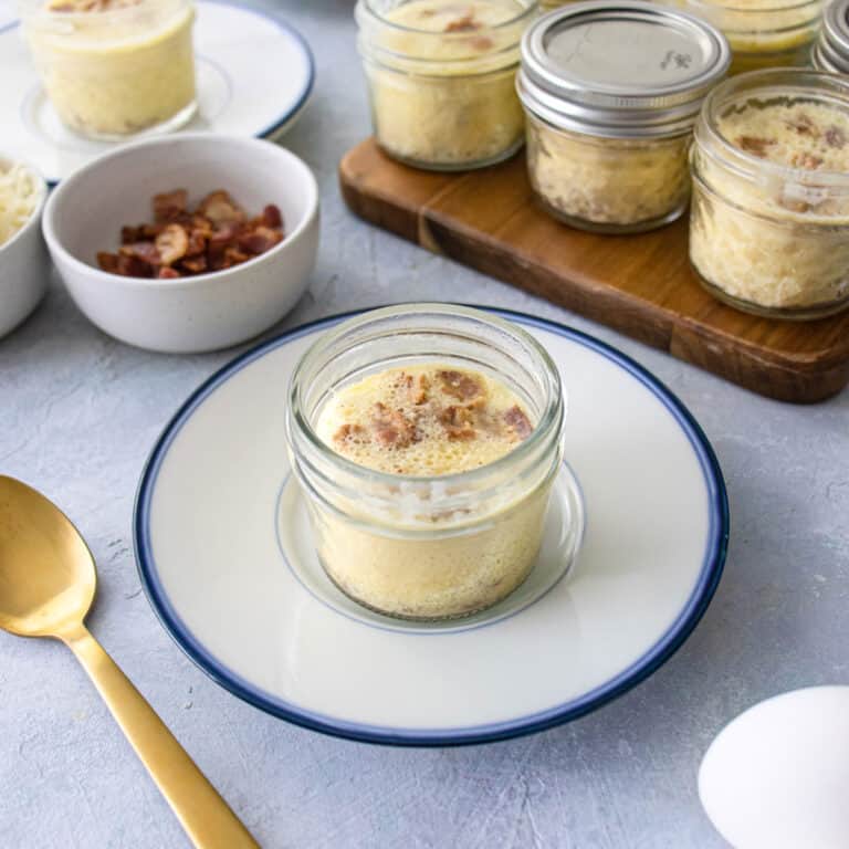 A small mason jar on a blue plate full of an egg bite. More mason jars in he background with a gold spoon and some bacon.