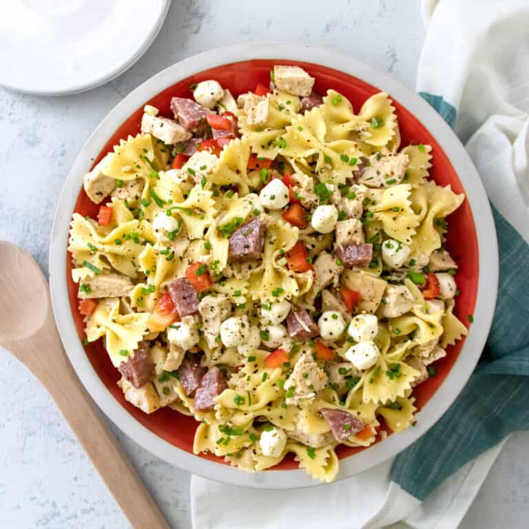 chicken bowtie pasta salad in a red serving bowl with a napkin, white plate and wooden spoon next to it.