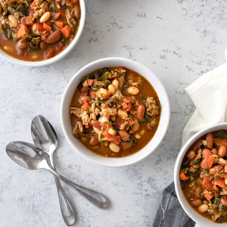3 small white bowls of 3 Bean Turkey and Kale Soup with 2 spoons laying next to it.