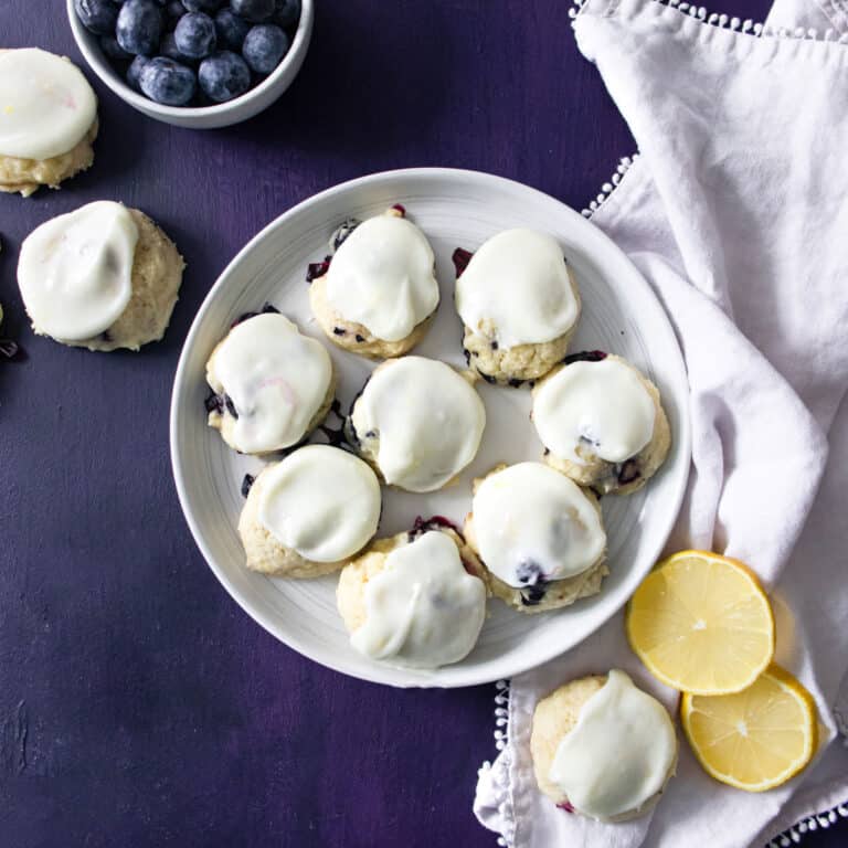 lemon and blueberry cookies on a white plate with cookies, lemon slices, and a bowl of blueberries next to them.