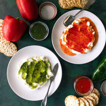 2 plates of green and red pepper jelly served over cream cheese with crackers and peppers on the table.