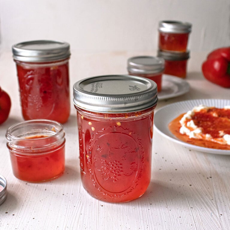Mason jars of red pepper jelly on a table with it served over cream cheese in the background.