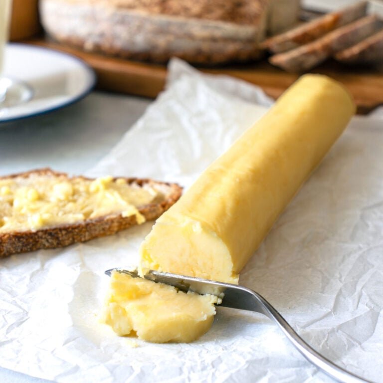A knife cutting into homemade butter with a loaf of sourdough in the background.