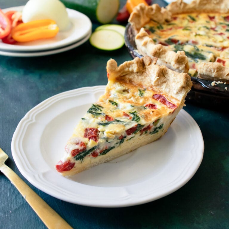 A slice of mixed veggie quiche on a white plate with the rest of it in the background.