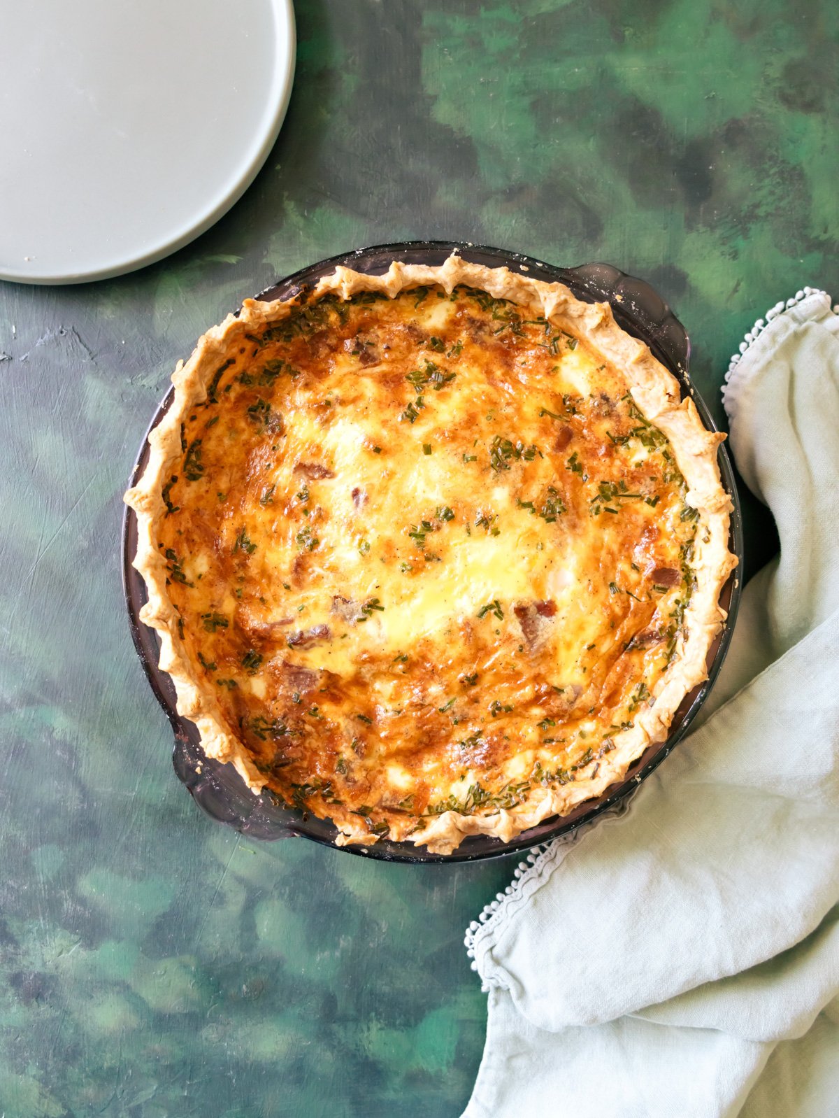 Crispy Bacon Onion Gruyere Breakfast Quiche on a green table with a napkin and plate around it. 