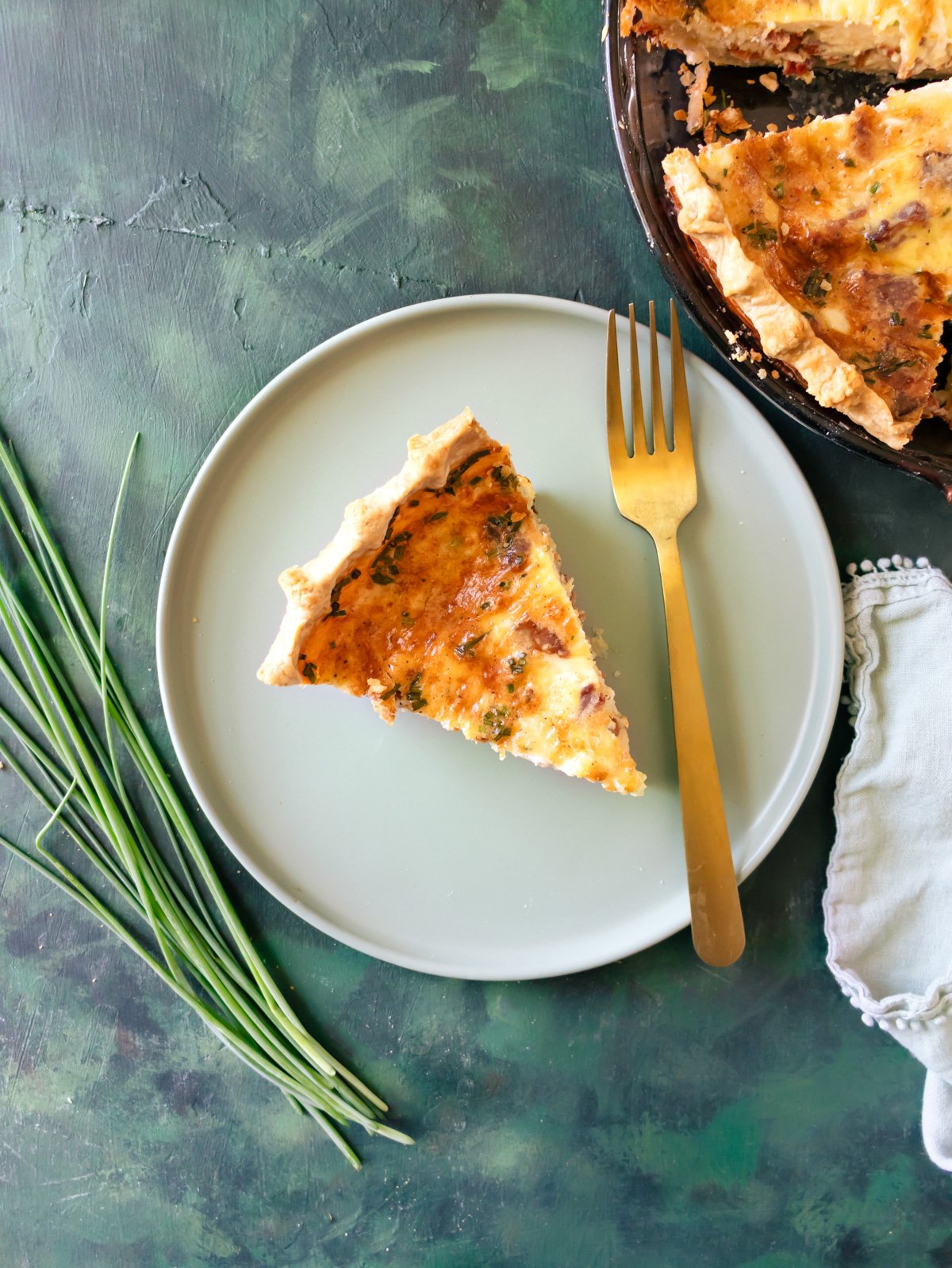 A slice of Crispy Bacon Onion Gruyere Breakfast Quiche on a green plates with a gold fork and the rest of the quiche behind it.