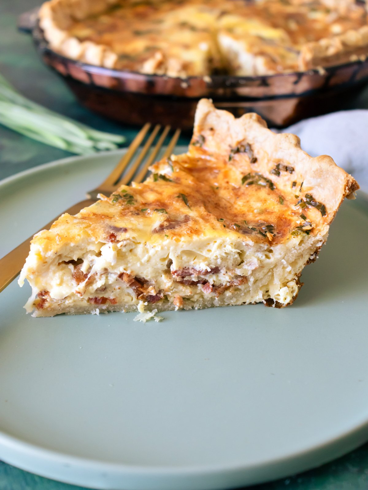 A slice of Crispy Bacon Onion Gruyere Breakfast Quiche on a green plates with a gold fork and the rest of the quiche behind it.