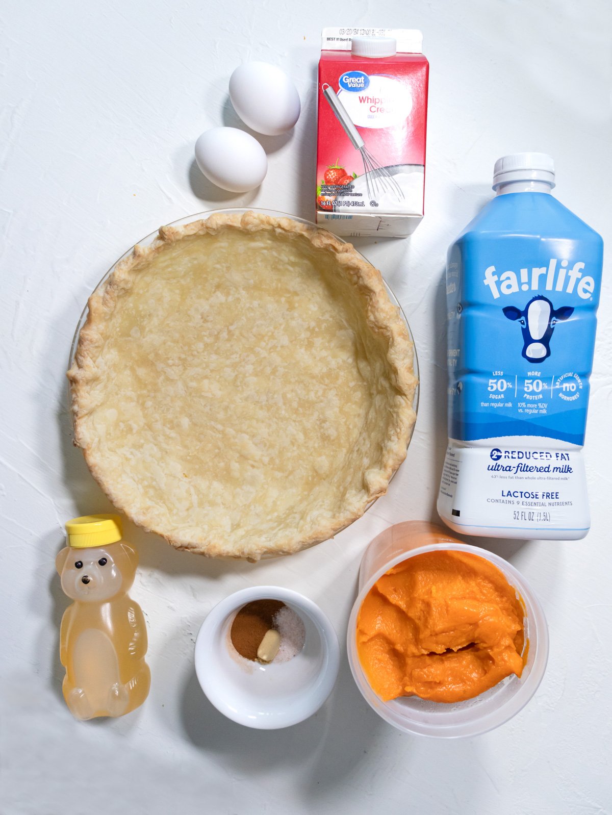 Eggs, whipping cream, milk, pie crust, sweet potato puree, spices and honey on a white table.