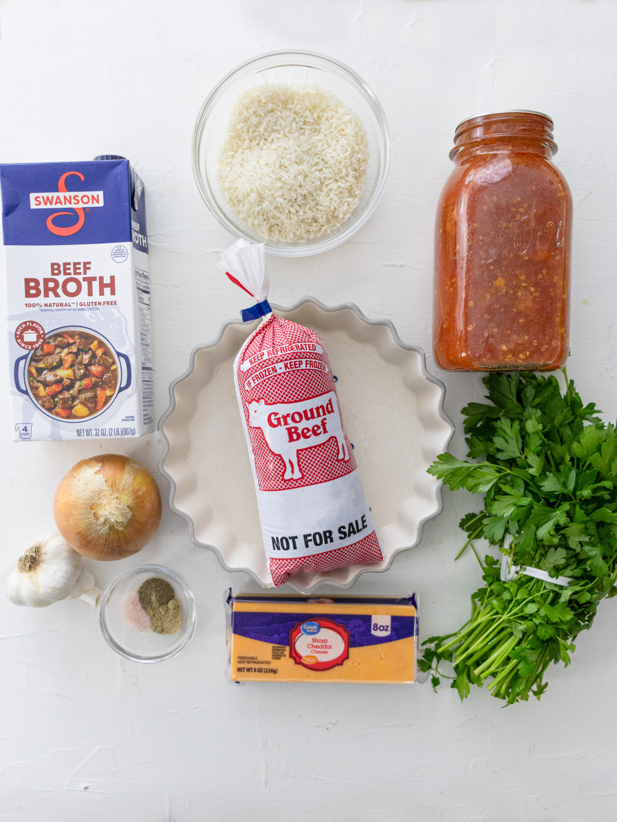 beef broth, ground beef, a bowl of rice, tomato sauce, fresh parsley, cheese, onion, garlic, and a bowl of spices on a white table.