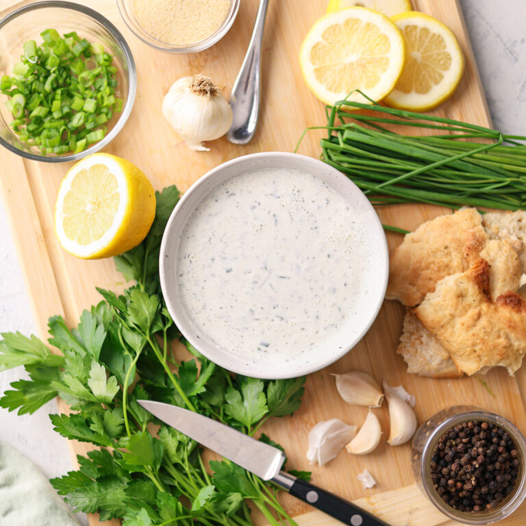 greek yogurt ranch dip on a bowl on a cutting board with veggies, herbs, lemon, and a knife.
