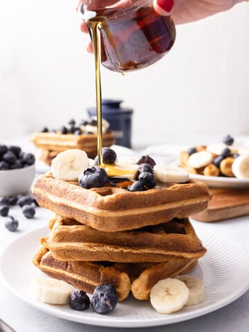 pouring maple syrup on top of a stack of waffles.