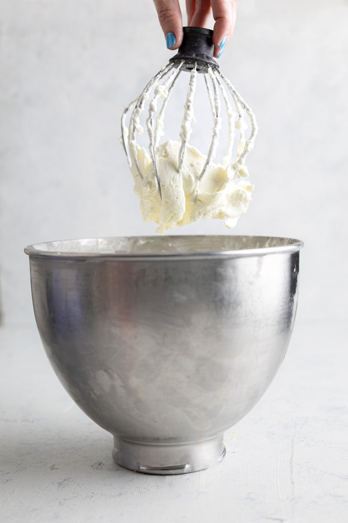 a hand holding a stand mixer whisk up above a bowl with whipped cream on top.