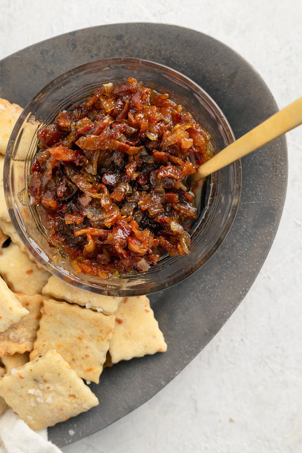 bacon jam in a clear bowl with a gold spoon in it and crackers on a platter next to it.