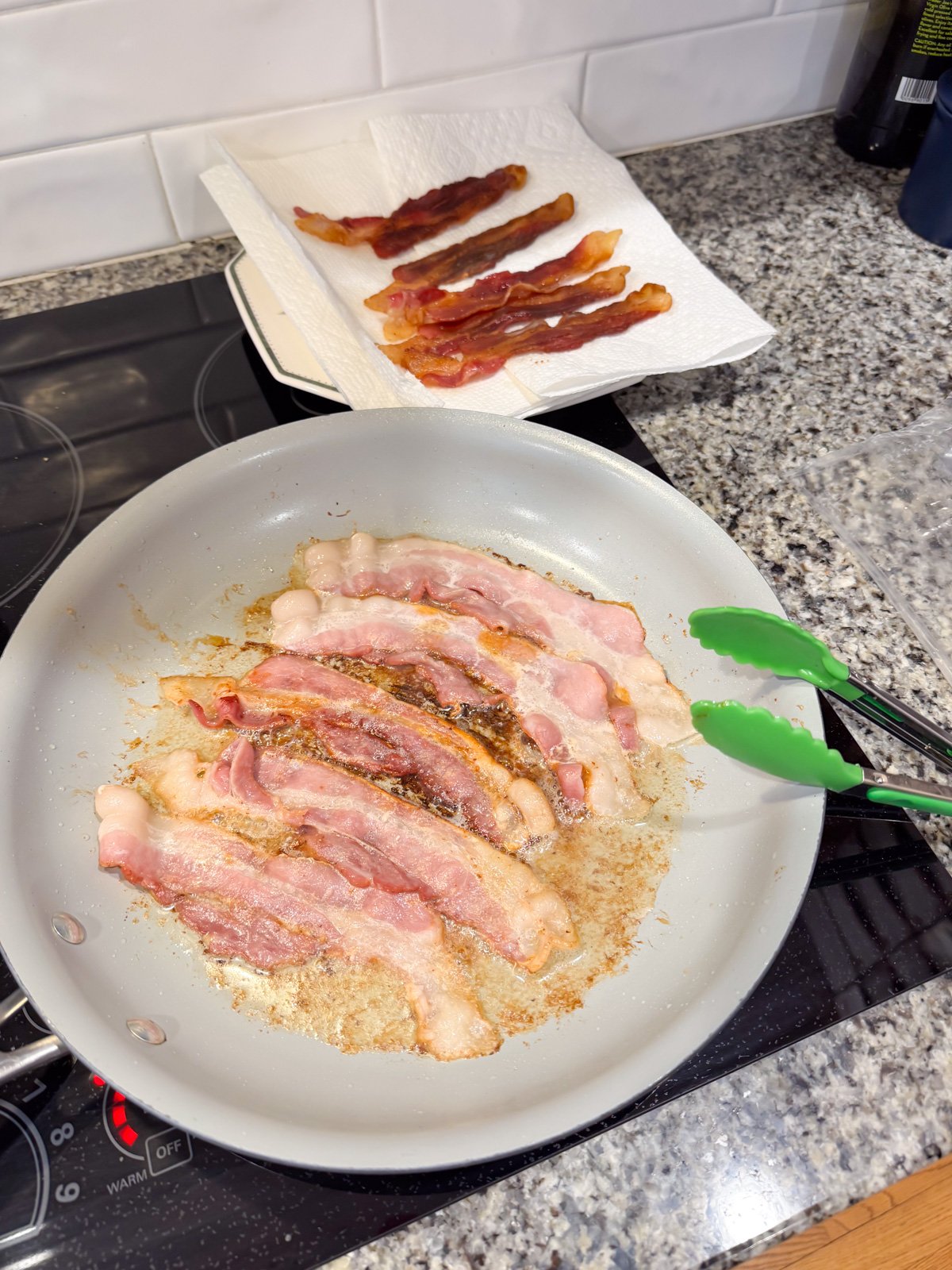 Cooking bacon on a skillet on the stove with tongs flipping them.