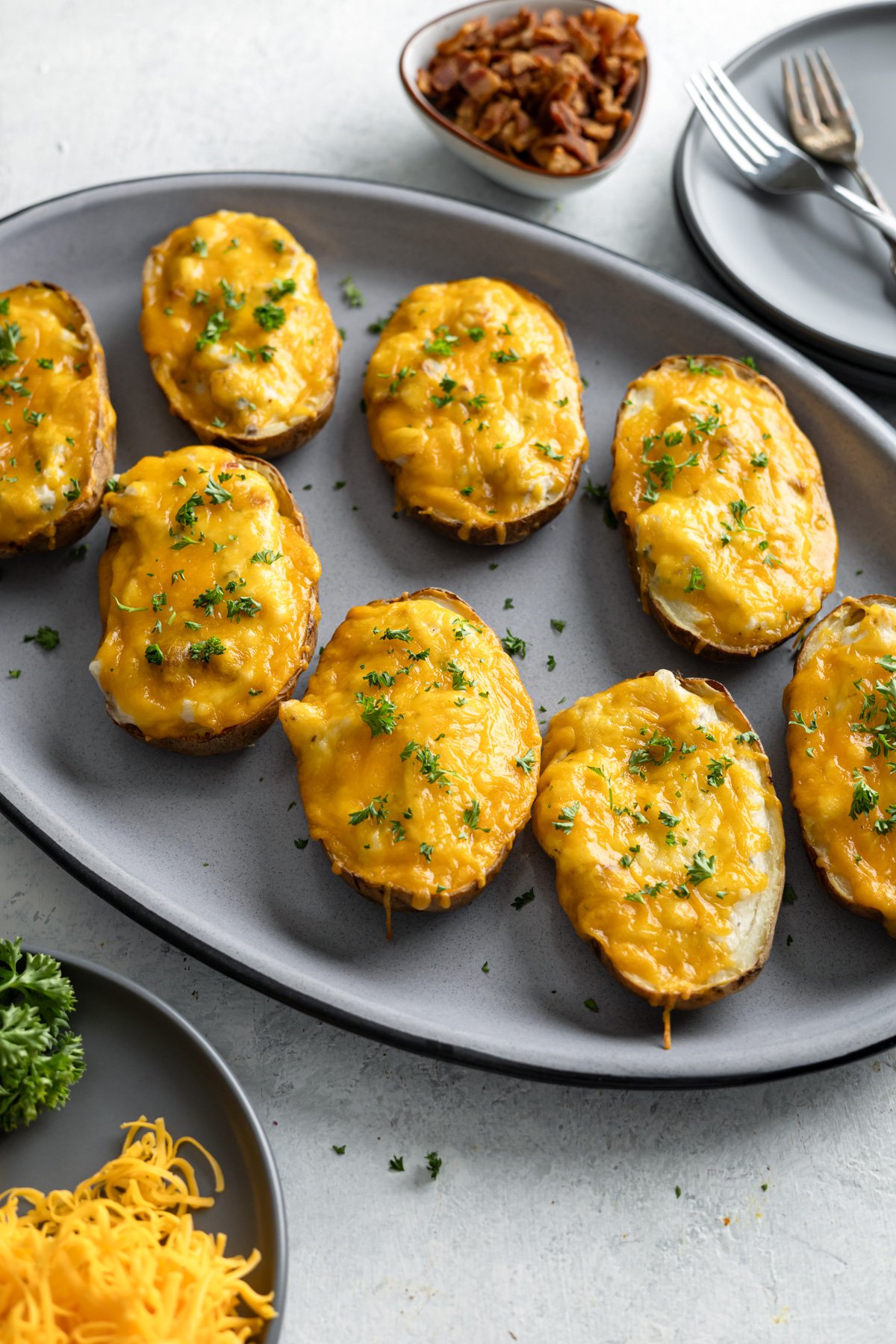 a platter of twice bake potatoes with plates and forks behind it and a small bowl of bacon bits.