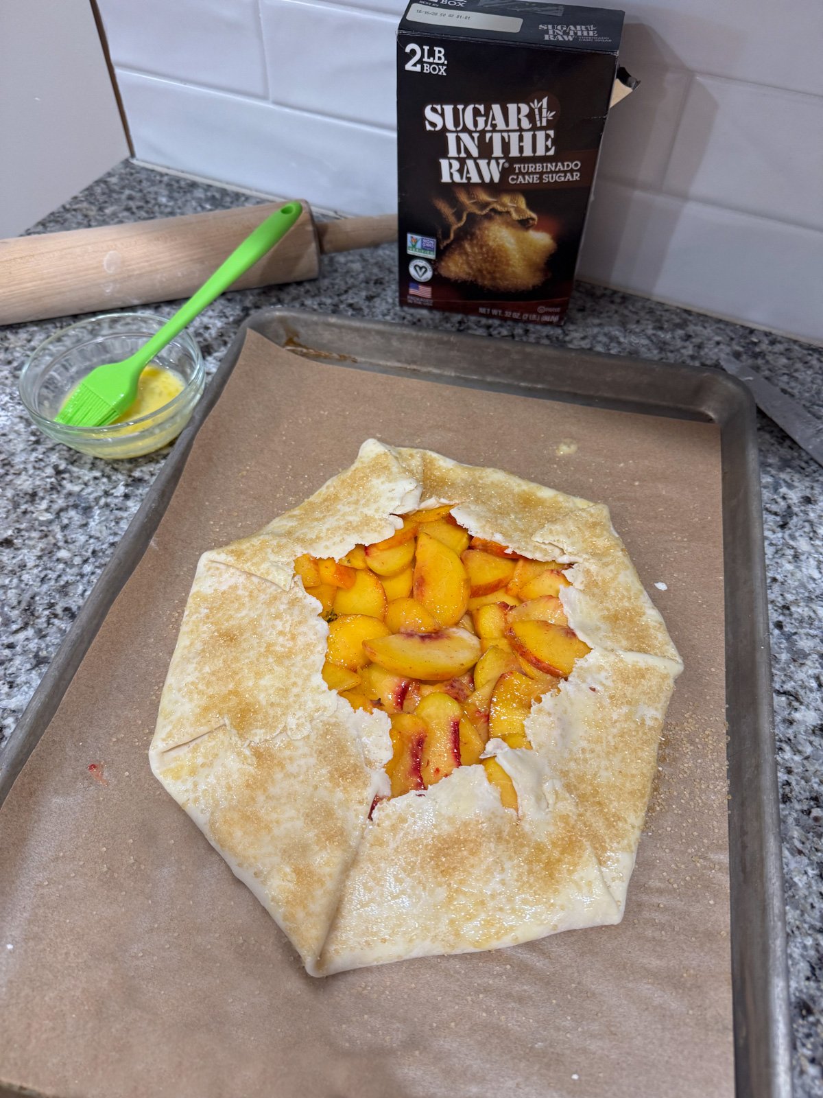 brushing an egg wash and sprinkling with sugar on a peach galette on a baking sheet before baking.