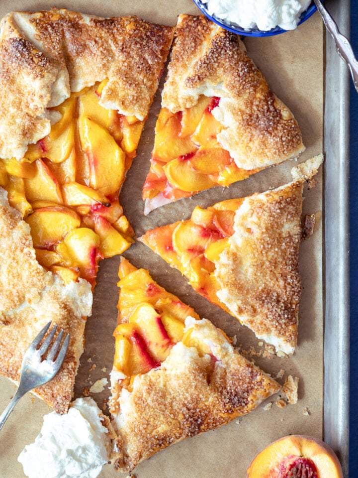 peach galette on a baking sheet with 3 pieces cut out and whipped cream next to it.