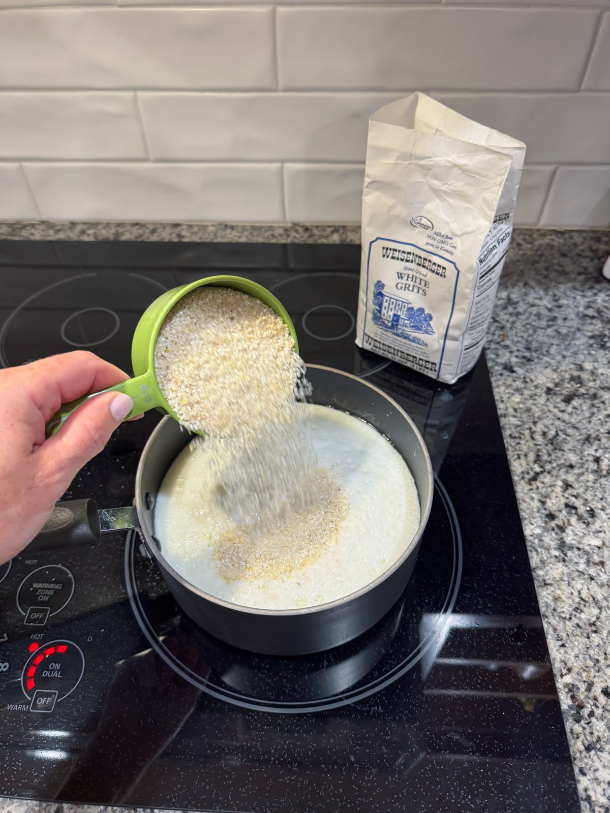 A hand pouring grits into a pot of shimmering milk on the stove.