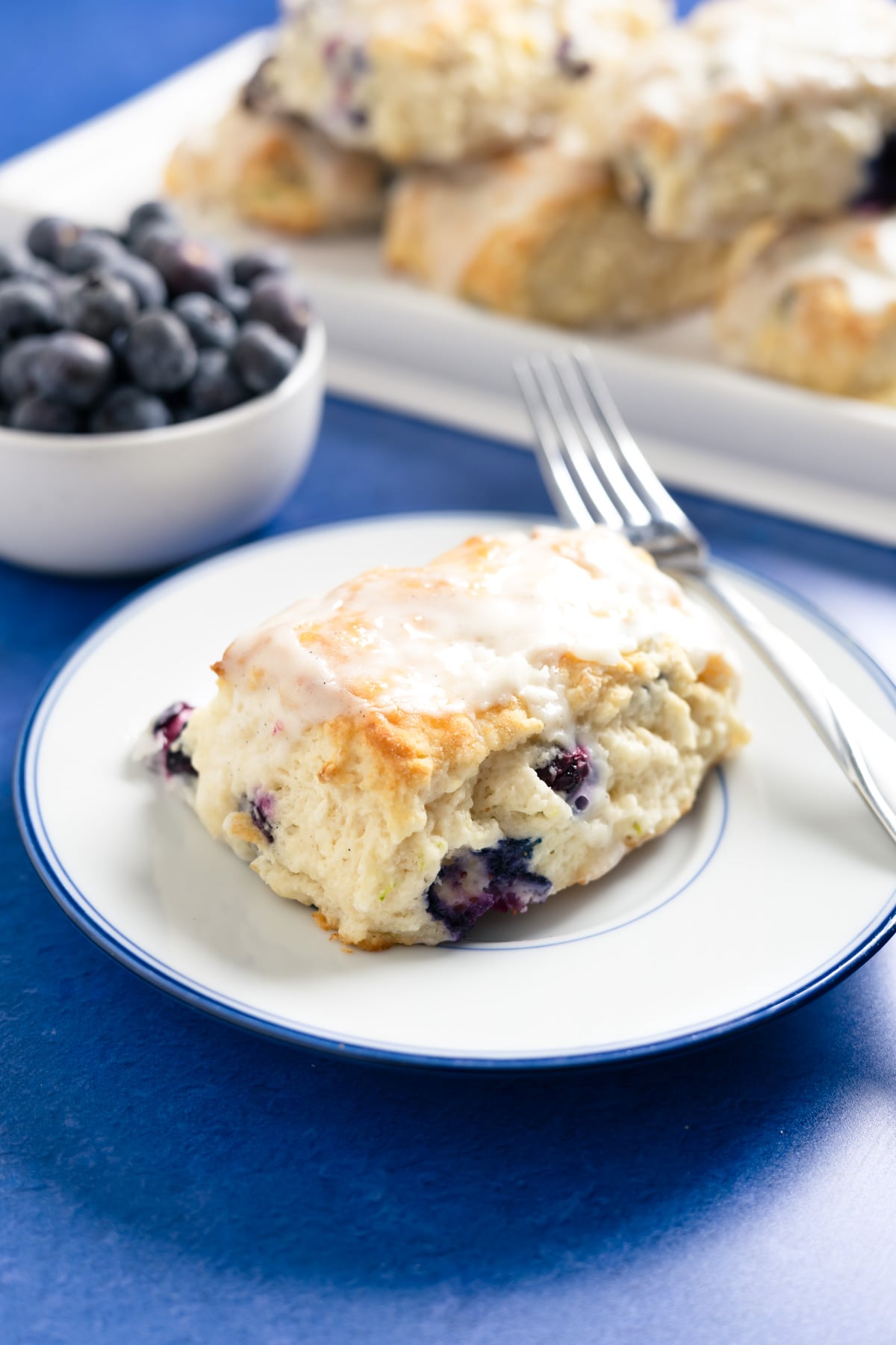 a small plate with a fork and blueberry buttermilk biscuit on it. a bowl of blueberries and a platter of biscuits in the background.