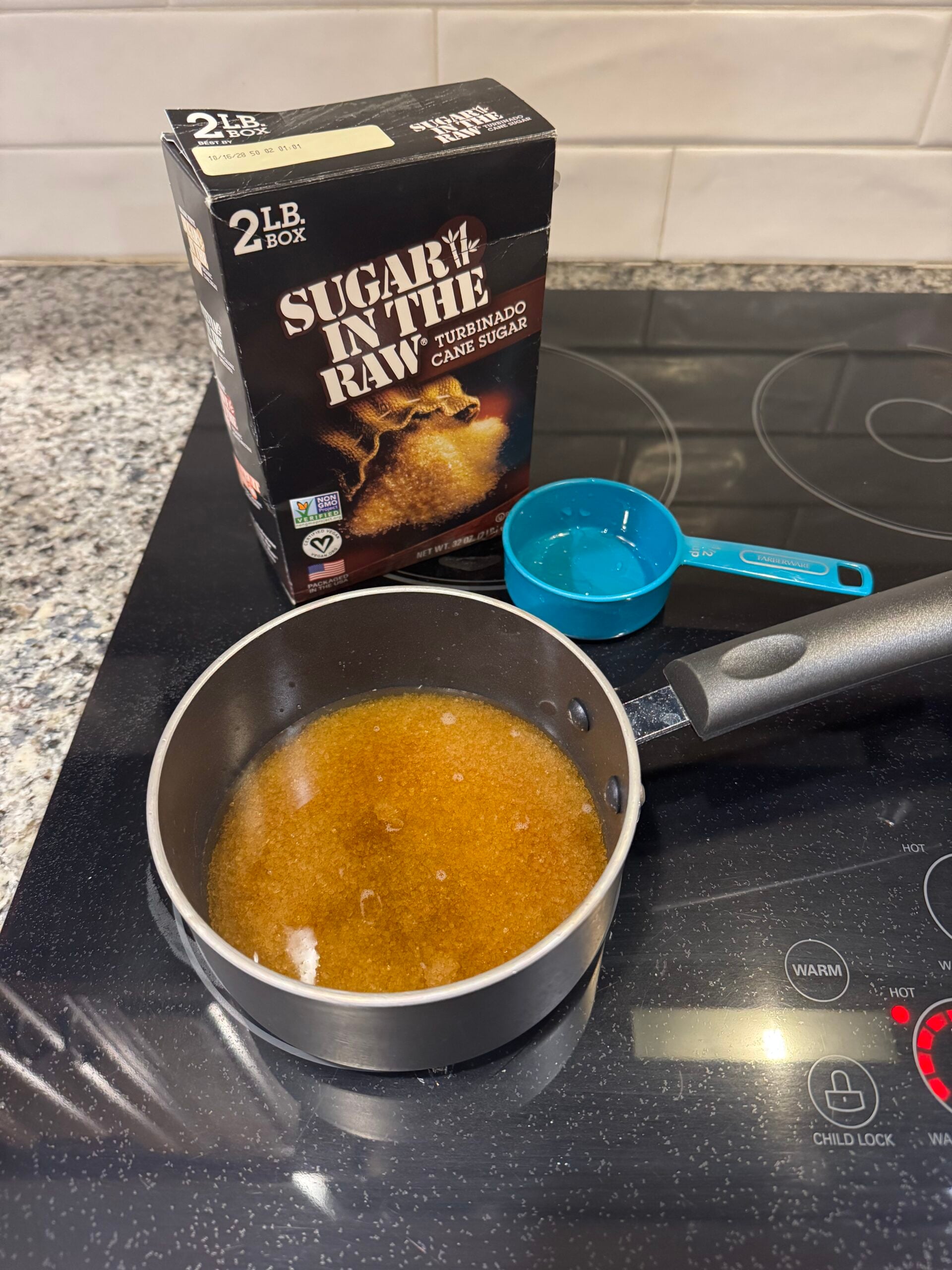 A saucepan with dissolved brown sugar for a large batch old fashioned sits on a stovetop. Behind it, a box of Sugar In The Raw turbinado cane sugar and a blue measuring cup rest on the counter.