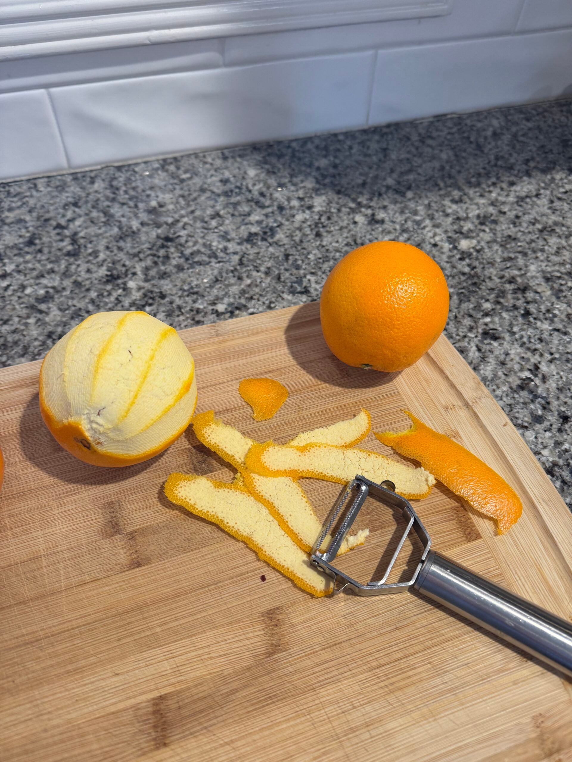 A peeled orange, an unpeeled orange, orange peels, and a metal peeler rest on a wooden cutting board-essentials for prepping a large batch old fashioned-on a granite countertop with a white tiled backsplash in the background.