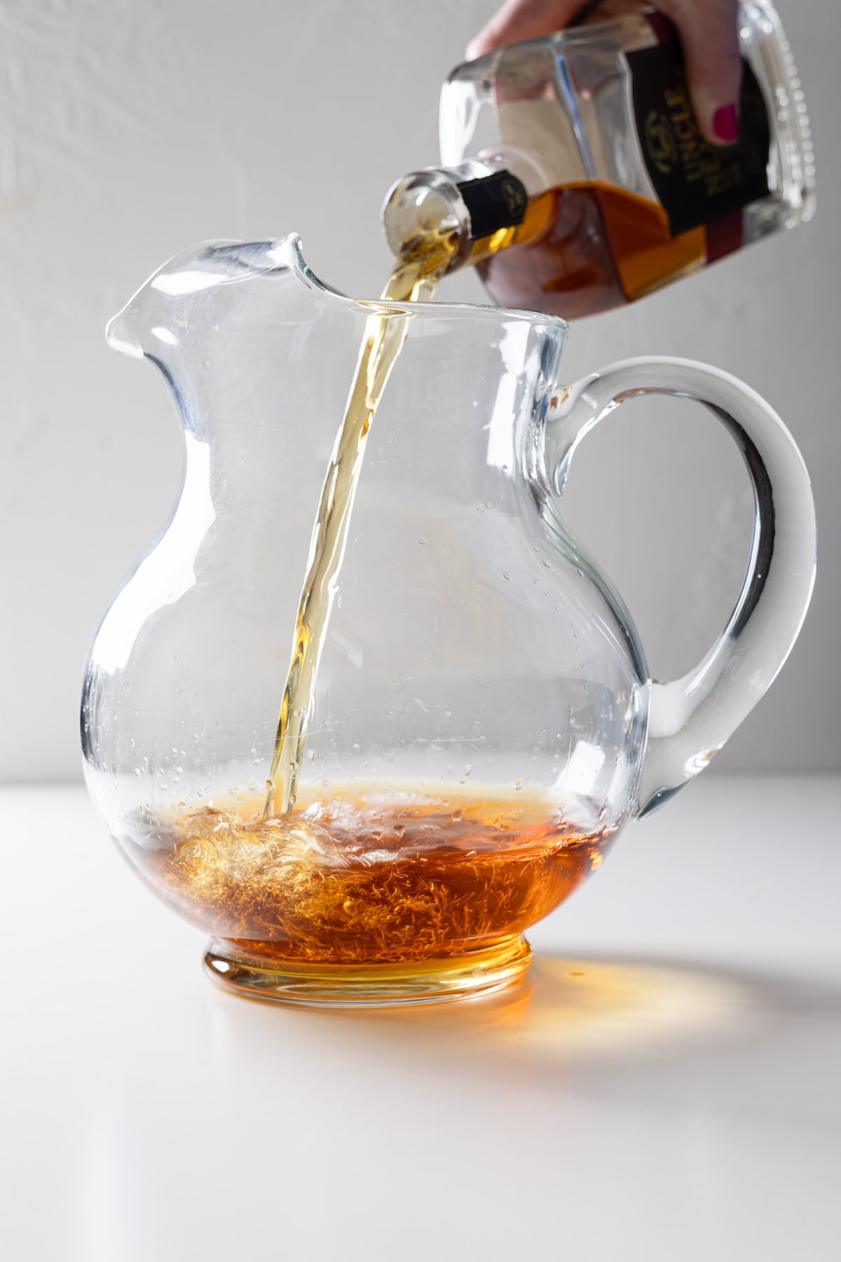A hand pours amber-colored liquid from a whiskey bottle into a clear glass pitcher on a white surface, preparing a large batch Old Fashioned as the liquid swirls at the bottom.