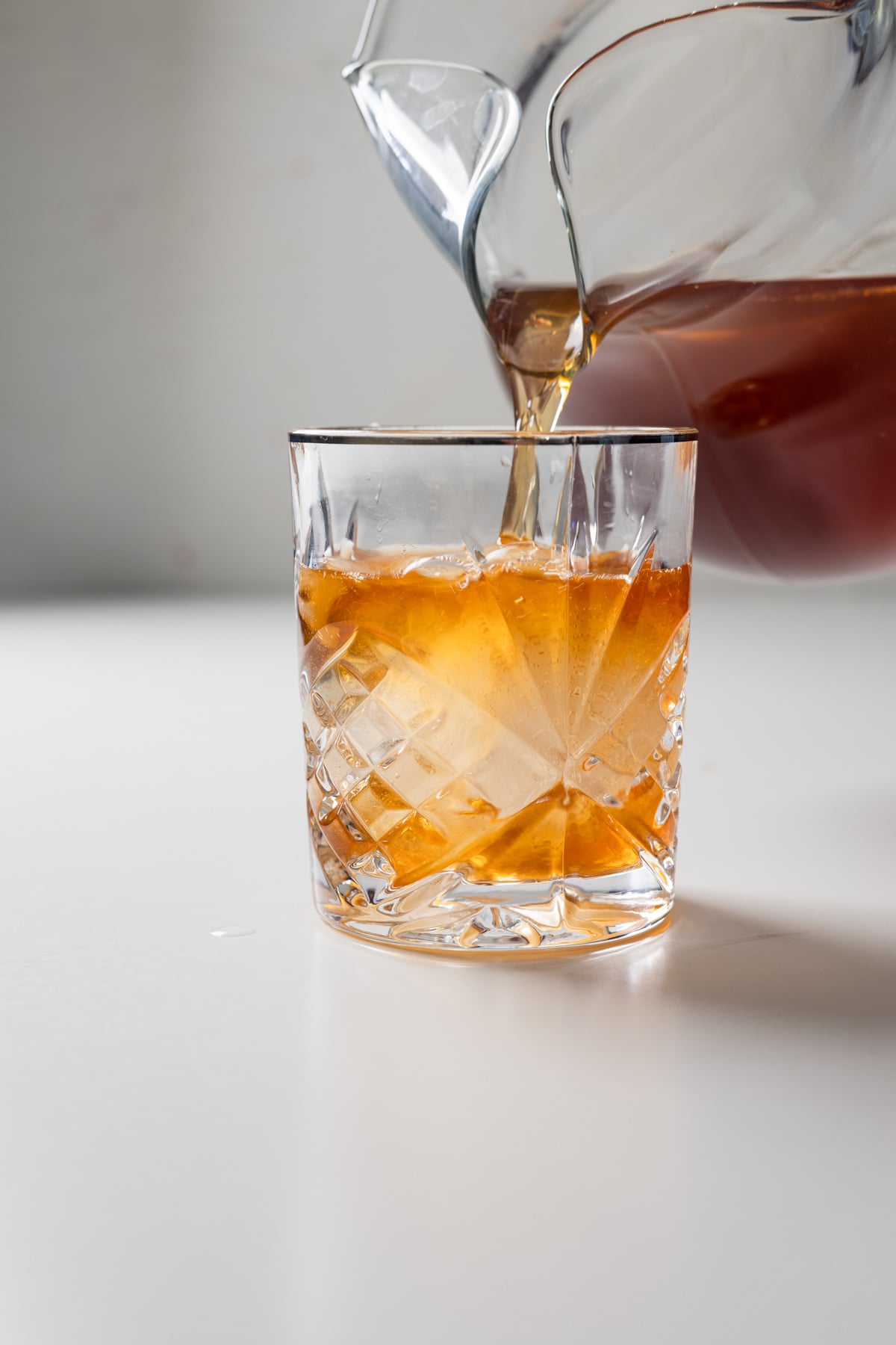 A glass pitcher pouring a large batch old fashioned, its amber hue cascading over ice cubes into a faceted glass tumbler on a white surface.