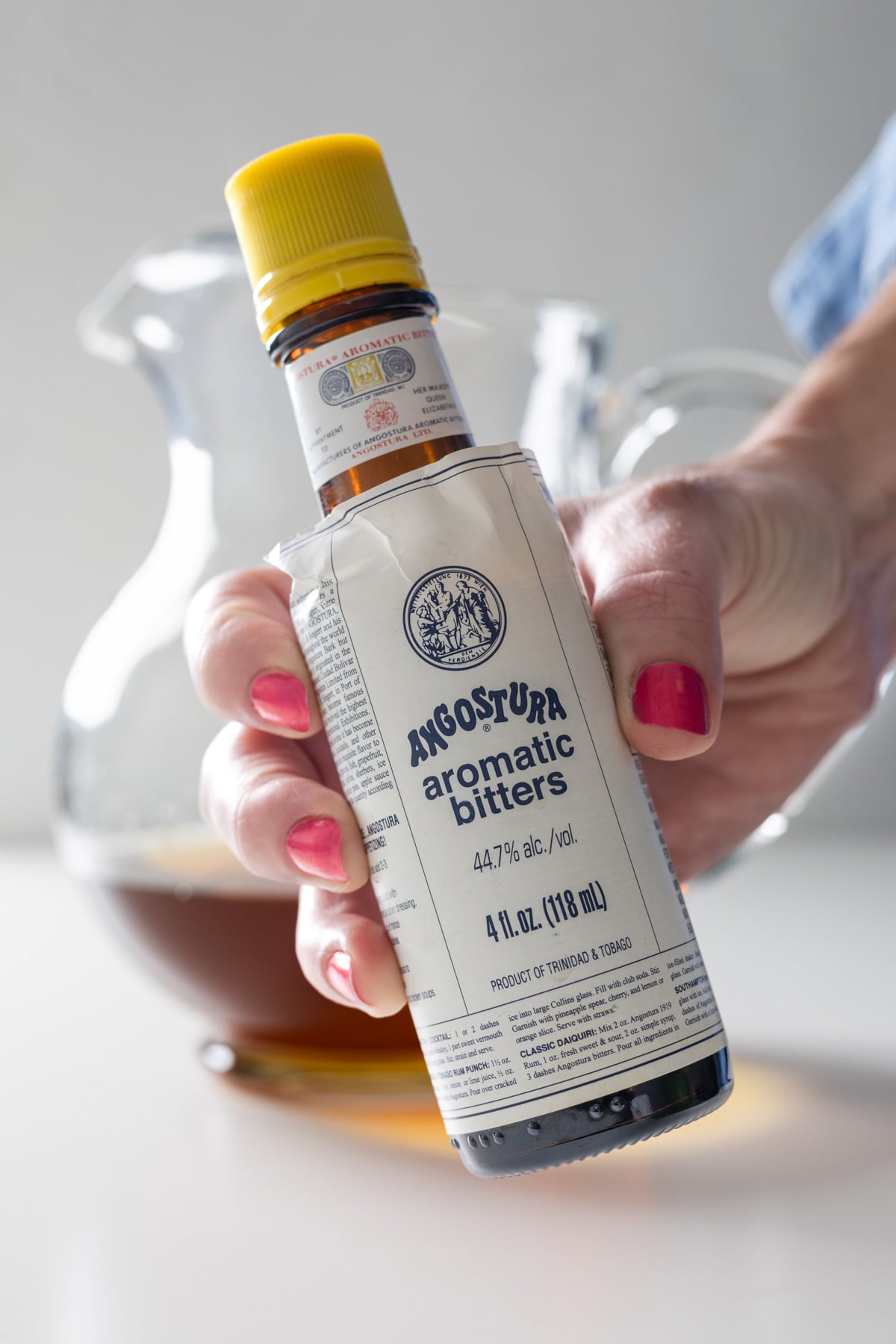 A hand with red-painted nails holds a bottle of Angostura aromatic bitters in front of a glass pitcher containing a brown liquid, ready for mixing a large batch old fashioned.